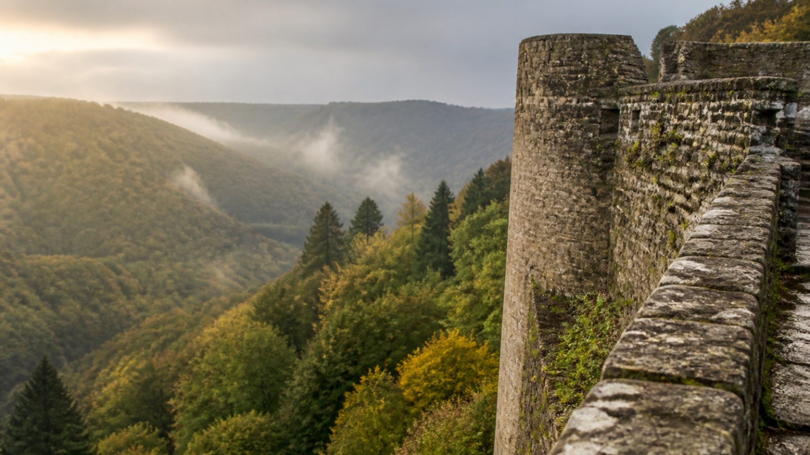 Weathered stone castle walls above a wooded Ardennes valley under soft autumn light.