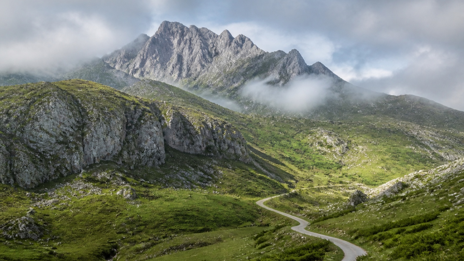 Layered limestone peaks above a green Cantabrian valley with a winding road and low Atlantic cloud.